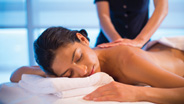 a woman lying on her front, relaxed with her eyes closed. a masseuse can be seen giving her a back massage.